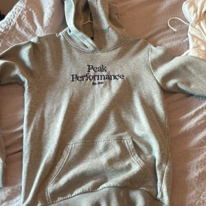 Grå hoodie från Peak Performance - Säljer en grå hoodie från Peak Performance med broderad logga på bröstet. Tröjan har huva, magficka och ribbade muddar vid ärmslut och nederkant. Perfekt för chill eller träning, riktigt skön och stilren.