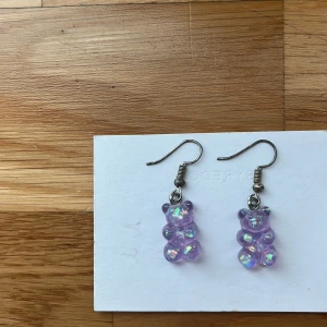Lila glittriga björnörhängen - Supergulliga örhängen med hängande björnar i lila transparent plast med glittriga detaljer. Perfekt för dig som vill ha något lekfullt och unikt i öronen. Krokarna är i metall och björnarna har en skimrande effekt som fångar ljuset. Jag själv är nickelallergiker och har inte haft några problem. 