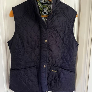 Quiltad Barbour väst med blommigt foder - Mörkblå quiltad väst från Barbour med snyggt blommigt foder i grönt, blått och vitt. Västen har två stora fickor framtill, dragkedja och tryckknappar. Perfekt för lager-på-lager och har en klassisk, tidlös look.