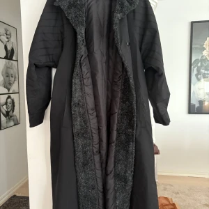 Svart lång kappa med fuskpäls från Glen - Lång svart kappa från Glen med bred grå fuskpäls längs krage och insida. Oversized passform, quiltade ärmar och knäppning framtill. Tillverkad i polyester och bomull, perfekt för kalla dagar när du vill ha en cool och mysig vibe. Vet ej strl men gissar på L 