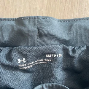 Grå träningsshorts från Under Armour - Snygga grå träningsshorts från Under Armour med elastisk midja och diskreta svarta paneler på sidorna. Shortsen är tillverkade i lätt polyester som andas och har en loose passform för maximal rörelsefrihet. Perfekta för gymmet eller löprundan.