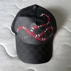 Svart Gucci keps med ormtryck - Snygg svart keps från Gucci med ikoniskt GG-mönster och en röd-vit orm tryckt på framsidan. Kepsen har mesh på baksidan och justerbar rem med metallspänne. Perfekt för dig som vill sticka ut med en lyxig accessoar.