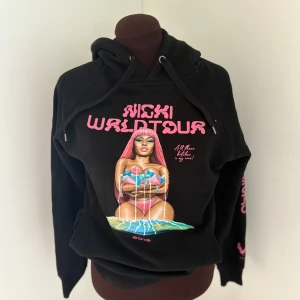 Nicki WRLDTOUR svart hoodie - Svart hoodie med färgglad Nicki Minaj WRLDTOUR-tryck framtill och rosa text på ärmen. Tröjan har huva med snörning och coolt grafiskt motiv i rosa, blått och grönt. Perfekt för dig som vill sticka ut och visa din fandom.