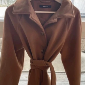 Brun kappa från Gina Tricot med bälte - Snygg brun kappa från Gina Tricot med klassisk krage och knappar framtill. Kappan har ett matchande bälte i midjan som ger en clean och stilren look. Perfekt för dig som gillar en tidlös vibe och vill hålla dig varm under kyliga dagar.