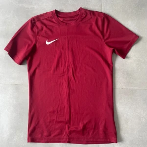 Vinröd Nike Dri-FIT t-shirt slim fit - Snygg vinröd t-shirt från Nike med klassisk vit swoosh på bröstet. Modellen är slim fit och tillverkad i Dri-FIT-material som andas, perfekt för träning eller sport. T-shirten har rund halsringning och korta ärmar för en sportig vibe.