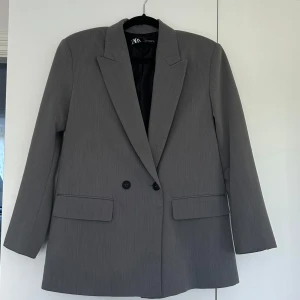 Grå kavaj från Zara - Stilren grå kavaj från Zara med klassisk krage och två knappar framtill. Rak passform och långa ärmar. Väldigt fint skick!