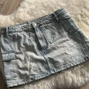 Ljusblå jeanskjol från Zara - Säljer en ljusblå jeanskjol från Zara i storlek L. Kjolen har klassiska fickor fram och bak, samt coola sidfickor med lock. Den är kort och har bälteshällor i midjan. Perfekt för dig som gillar en chill och trendig denimlook.
