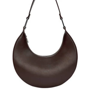 Mörkbrun axelväska i skinnimitation - Säljer en snygg mörkbrun axelväska i halvmåneformad design. Får plats med mobil, laddare, solglasögon, läppstift, nycklar & korthållare. Så väldigt rymlig! Endast använd 1 gång! Finns inte att köpa längre!