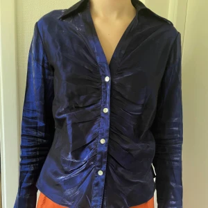 Mörkblå glansig skjorta med knappar - Snygg mörkblå skjorta med glansig finish och markerad rynkning längs knappraden. Skjortan har lång ärm, klassisk krage och figurnära passform.