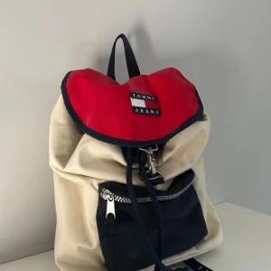 Tommy Jeans ryggsäck i rött och beige - Snygg ryggsäck från Tommy Jeans i beige canvas med röd lock och mörkblå detaljer. Väskan har ett stort huvudfack med dragsko, ytterficka med dragkedja och justerbara axelband. Klassisk logga framtill och coolt band med text. Perfekt för skolan eller stan. Priset är förhandlingsbart!