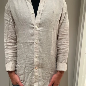 Beige linneskjorta med knappar - Snygg beige skjorta i linne med klassisk krage och knäppning framtill. Långärmad modell med avslappnad passform och diskret broderad logga på bröstet. Perfekt för en stilren och chill look. Köptes nyligen, nypris 1699kr. Måste rensa skjortorna tyvärr. S passar också M