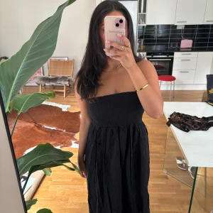 Svart axelbandslös långklänning / maxikjol - Tänkt att kunna bäras som maxi kjol eller klänning. Säljer en svart axelbandslös långklänning med smockad överdel och rynkad kjol. Klänningen har en enkel och stilren look, perfekt för dig som gillar minimalistisk stil. HELT NY