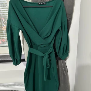 Grön omlottklänning från Boohoo - Snygg grön omlottklänning från Boohoo med v-ringad hals och knytband i midjan. Klänningen har trekvartsärmar med puff och är tillverkad i ett mjukt syntetmaterial. Perfekt för dig som gillar stilrena och trendiga plagg med lite extra detaljer.