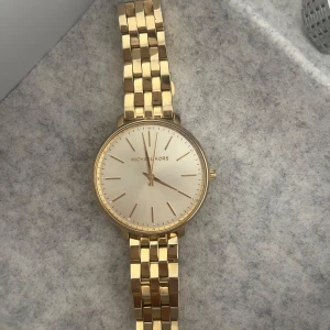 Guldfärgad klocka från Michael Kors - Stilren guldfärgad klocka från Michael Kors med klassisk rund urtavla och länkarmband i metall. Urtavlan är ljus med minimalistiska markeringar och diskret logga. Perfekt accessoar för dig som gillar en clean och elegant look.