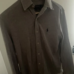 Grå skjorta från Polo Ralph Lauren XS - Snygg grå skjorta från Polo Ralph Lauren i storlek XS. Skjortan är i ett lätt och mjukt bomullsmaterial, har klassisk krage, knäppning framtill och broderad logga på bröstet. Perfekt för dig som gillar stilren och enkel design.