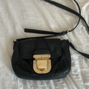 Svart axelväska från Michael Kors - Snygg svart axelväska från Michael Kors (gammal kollektion) i äkta skinn med justerbar rem och guldiga metalldetaljer. Väskan har ett stilrent spänne framtill med logga och ett klassiskt, tidlöst utseende. Perfekt storlek för det viktigaste och passar till många outfits. Säljes pågrund av att den ej används mer. Note to self, spännet framtill av väskan är lite repat därav kan priset diskuteras. Väskan intill har 3 fack, ett stort imitten och två lite mindre på vardera sida. 