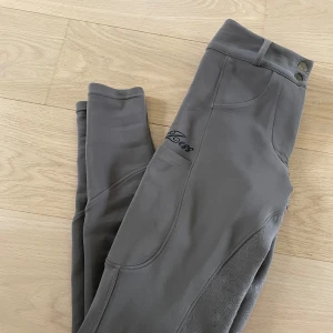 Grå helskodda ridbyxor från Zaczess  - Snygga grå ridbyxor från Zaczess med hög midja, silikonprint på insidan av benen för extra grepp och svarta detaljer vid bakfickorna. Byxorna har två knappar i midjan, bälteshällor och praktiska fickor på sidorna. Perfekta för ridning och riktigt bekväma. Gylfen har tyvärr gått sönder men det går att sätta i den enkelt, fullt användnings bara!