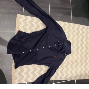 Mörkblå skjorta från Ralph Lauren - Klassisk mörkblå skjorta från Ralph Lauren med röd broderad logga på bröstet. Skjortan har lång ärm, vita knappar och en tidlös passform. Perfekt för dig som gillar stilrena och snygga plagg med ikonisk detalj.