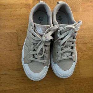 Beige sneakers från Adidas - Snygga beige sneakers från Adidas med klassiska tre ränder på sidan och vit sula. Skorna har snörning och är tillverkade i textil med detaljer i syntet. Perfekta för en clean och stilren look.