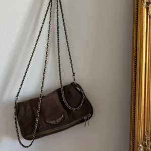 Brun mocka Zadig Väska - Snygg brun axelväska i mocka med silverfärgad kedjerem och dragkedja. Väskan har en cool vingdetalj framtill med glittriga stenar och en praktisk ficka med dragkedja på baksidan. Perfekt storlek för det viktigaste och ger en edgy vibe till din outfit.