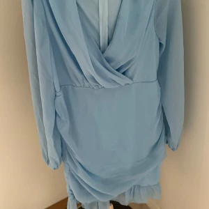 Ljusblå omlottklänning med volang - Säljer en ljusblå kortklänning med omlottdetalj och volang nedtill. Klänningen har långärmade ärmar och är gjord i ett lätt, flowy material som ger en snygg siluett. Perfekt för dig som gillar romantiska och trendiga plagg.