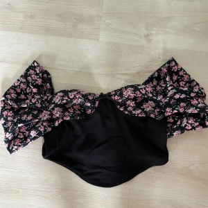 Svart tröja med blommiga volanger - Supersnygg offshoulder i svart med stora volanger över axlarna i blommigt mönster med rosa och gröna detaljer. Modellen är axelbandslös och har en cool, statement look som verkligen sticker ut. Perfekt för dig som vill ha något unikt och trendigt i sommar.