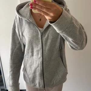 Grå hoodie med dragkedja från 157 - Säljer en ljusgrå hoodie från 157 i storlek XS. Tröjan har huva med snörning, praktiska fickor framtill och hel dragkedja. Mjuk insida i fleece och långärmad passform, perfekt för chill dagar eller när du vill vara extra comfy. (Stor i storleken)