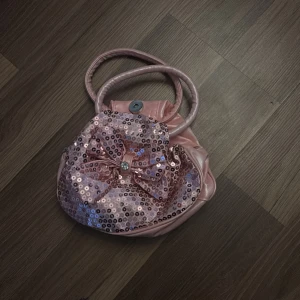 Rosa handväska med paljetter och rosett - Söt rosa handväska i syntetmaterial med två handtag. Framsidan är dekorerad med glittriga paljetter och en stor rosett med en glittrig sten i mitten. Väskan stängs med tryckknapp och har en rundad form som ger en gullig look.