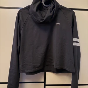 Svart croppad hoodie från Stronger - Svart croppad hoodie från Stronger med bred krage och vita ränder på ena ärmen. Tröjan har lång ärm och är gjord i mjuk bomull, perfekt för chill eller träning. Diskret logga på bröstet och cool, sportig vibe. 💕 orginalpris 699kr, tar emot prisförslag! 🤩