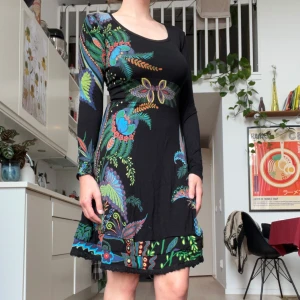 Svart blommig kortklänning med lång ärm från Desigual - Säljer en svart kortklänning med färgglatt blommigt mönster i blått, grönt, rött och lila. Klänningen har lång ärm, rund halsringning och en figurnära passform. Materialet är mjukt och stretchigt, perfekt för dig som gillar unika prints och vill sticka ut.