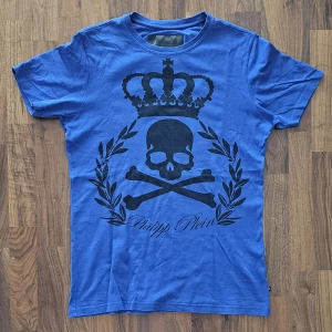 Blå Philipp Plein t-shirt med döskalle - Snygg blå t-shirt från Philipp Plein med stort svart tryck av döskalle, korslagda ben, krona och lagerkrans på bröstet. Klassisk passform, kortärmad och i mjuk bomull. Märkets logga i metall på ryggen ger extra edge till looken.