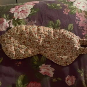 Blommig sovmask i bomull! - Supersöt sovmask med blommigt mönster i pastelliga toner av rosa, orange och grönt. Masken har en mjuk, vadderad känsla och ett elastiskt band baktill för skön passform. Perfekt för dig som vill sova extra gott och samtidigt ha en gullig accessoar.