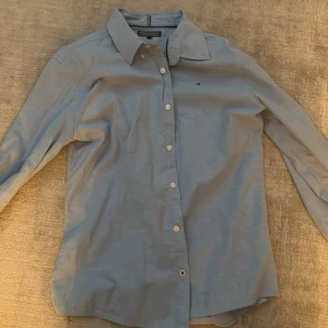 Ljusblå skjorta från Tommy Hilfiger - Snygg ljusblå skjorta från Tommy Hilfiger med klassisk krage och knäppning framtill. Skjortan har lång ärm och är tillverkad i mjuk bomull. Liten broderad logga på bröstet ger en clean och stilren look. Öppen för prisförslag 
