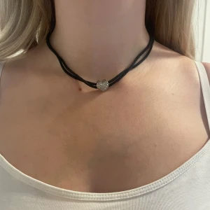 Svart choker med glittrigt hjärta - Snygg svart choker med dubbla band och ett silverfärgat hjärthänge med silverbubblor. Halsbandet har justerbar kedja och karbinlås. Perfekt för att ge din outfit en cool och trendig touch.