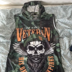 Tränings line - Säljer en ärmlös hoodie i camouflage med stora tryck av dödskallar och texten 'Veteran' och 'Liberty or death'. Tröjan har huva och coola militärinspirerade detaljer i grönt, svart och orange. Perfekt för dig som gillar streetwear med attityd.