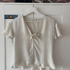 Vit blus med knyt från Vila - Supersöt vit blus från Vila med v-ringning, knyt framtill och små knappar längs hela framsidan. Blusen har korta ärmar och volangkant nertill som ger en romantisk vibe. Perfekt till jeans eller kjol för en fräsch look. Passar jättebra till sommaren. Den ser ganska bred ut på bild men det är det inte, den sitter löst och fint.🥰🥰