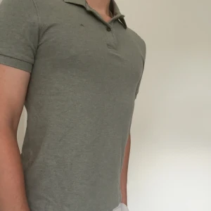 Grå pikétröja från J.Lindeberg - Snygg grå pikétröja från J.Lindeberg med klassisk krage och knappslå. T-shirten har korta ärmar och en stilren, enkel design utan mönster. Perfekt för dig som gillar en clean och sportig look. Materialet är mjuk bomull som känns skönt mot huden.