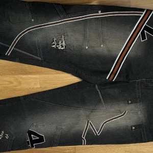 Svarta baggy jeans med broderier - Säljer ett par svarta baggy jeans med coola broderade detaljer och kontrastfärgade ränder i vitt och brunt. Jeansen har flera fickor, slitningar och stora bokstavsbroderier som ger en unik streetkänsla. Perfekt för dig som gillar en avslappnad och edgy stil.