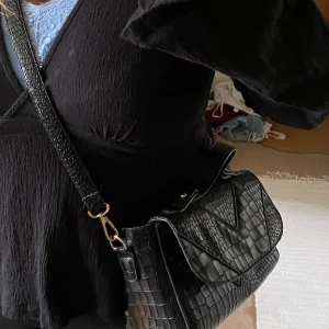 Svart axelremsväska - Snygg svart axelremsväska med avtagbar axelrem. Väskan är använd men ändå i bra skick. Man får plats med mycket i väskan. Mått: 24x18x9