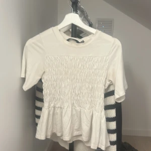 Vit blus med smock från Vero Moda - Supersöt vit blus från Vero Moda med smockad front och volangkant nedtill. Blusen har korta ärmar och rund halsringning. Tillverkad i mjuk bomull, perfekt för dig som gillar en feminin och chill stil.