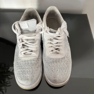 Nike Air Force 1 Flyknit vita sneakers - Nike Air Force 1 Flyknit i helvitt med klassisk Swoosh på sidan. Ovandelen är stickad i mesh för en lätt känsla och snygg textur, med vit snörning. Perfekta för dig som gillar stilrena och sportiga sneakers.