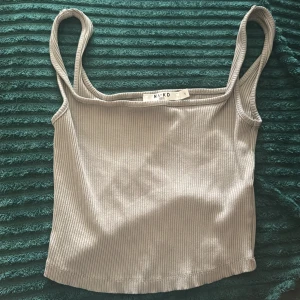 Ribbad grå linnetopp från NA-KD - Säljer en stilren grå ribbad linnetopp från NA-KD. Toppen är ärmlös med smala axelband och har en croppad passform. Perfekt för varma dagar eller att styla lager på lager. Enkel och snygg att matcha med jeans eller kjol.