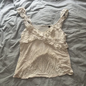 Vit linnetopp med volangdetaljer - Supersöt vit linnetopp med breda axelband och volangdetaljer längs ringningen och banden. Perfekt för varma dagar och ger en romantisk vibe till outfiten. Tunt och lätt material som känns luftigt.