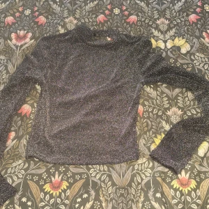 Glittrig svart topp från H&M Divided - Snygg svart glittrig topp från H&M Divided i storlek XS. Toppen har lång ärm, hög hals och en öppen rygg som ger en cool och trendig vibe. Perfekt för dig som vill sticka ut lite extra med ett glittrigt plagg.