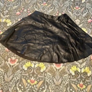 Svart skinnkjol med dragkedja - Snygg svart kort skinnkjol med lätt utsvängd form och guldig dragkedja bak. Kjolen har en clean look och passar perfekt till en edgy outfit. Materialet är glansigt och ger en cool vibe.