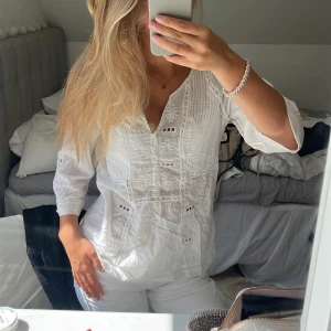 Vit blus med broderade detaljer - Säljer en luftig vit blus med broderade blommor och spetsdetaljer. Blusen har v-ringning, trekvartsärm och är tillverkad i tunn bomull. Köpt second hand. Klicka gärna på ”köp nu”!