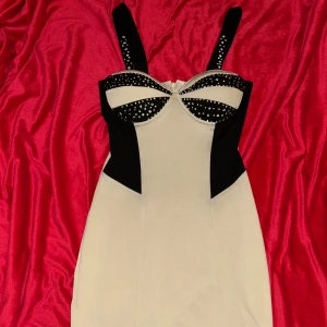 Beige och svart midi klänning  - Snygg beige och svart kort klänning med breda axelband. Klänningen har en figurnära passform och kontrastpaneler som ger en riktigt edgy look. Dragkedja bak för enkel påklädning. Perfekt för dig som gillar statement-plagg! Någon enstaka detalj (blingmärkena) har ramlat av, men inget som man lägger märke till om man inte verkligen kollar 