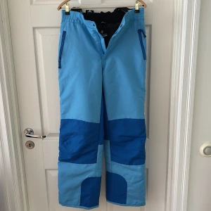 Blå täckbyxor med hängslen - Säljer ett par täckbyxor i två nyanser av blått med svarta och vita hängslen. Byxorna har dragkedjor vid fickorna, förstärkta knän och justerbara benslut. Perfekta för vinterns alla äventyr och håller dig varm och torr i snön. Barnstorlek!