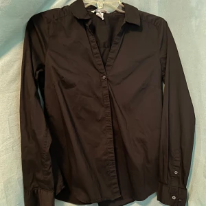 Svart klassisk skjorta  - En stilren svart skjorta med lång ärm och klassisk krage med viss urringning. Skjortan har knappar framtill och vid ärmsluten, vilket ger en clean och tidlös look. Perfekt för dig som gillar enkel och snygg stil. Materialet känns mjukt och bekvämt mot huden.