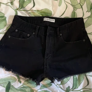 Svarta jeansshorts från Pull&Bear - Svarta jeansshorts från Pull&Bear med mid waist och fransiga kanter. Klassisk femficksmodell med slitna detaljer på bakfickorna för en edgy vibe. Perfekta till sommaren och lätta att matcha med allt. I storlek 36
